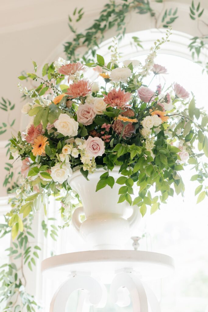 A large white vase holds a lush bouquet of pink, peach, yellow, and white flowers with abundant green leaves, set in front of a sunlit window framed by leafy vines—perfect for a progressive cocktail reception Atlanta style.