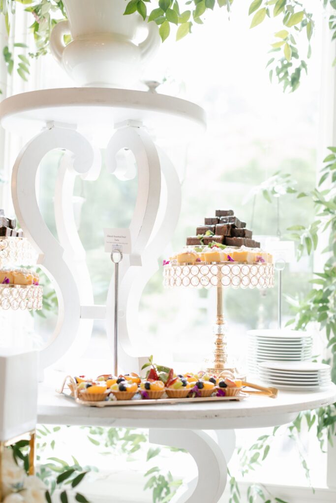 A bright, elegant dessert table for a progressive cocktail reception in Atlanta, with fruit tarts and brownies on gold stands, surrounded by stacked white plates and lush green foliage under soft natural light.