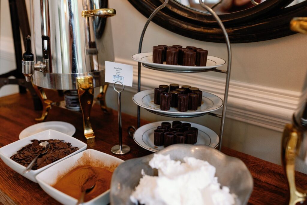 A hot beverage station perfect for a progressive cocktail reception in Atlanta, featuring a metal dispenser, a three-tiered tray of small brown cups, and bowls of whipped cream, chocolate shavings, and cocoa powder on a wooden table.