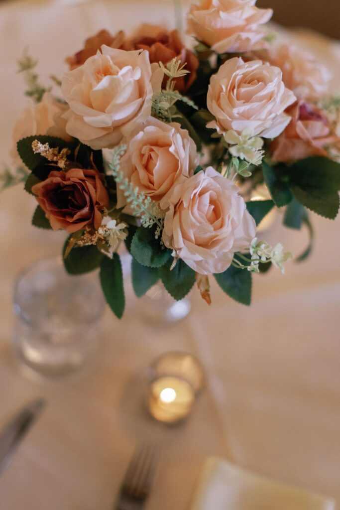 A bouquet of light pink and peach roses with greenery in a vase sits on a table, styled for a progressive cocktail reception Atlanta, surrounded by a glass of water, a lit tealight candle, and silverware on a cream-colored tablecloth.