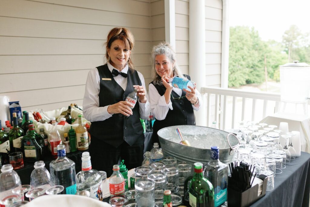 Two smiling bartenders in uniforms stand behind a bar set up for a progressive cocktail reception in Atlanta, preparing drinks with assorted liquor bottles, mixers, and glassware on an outdoor patio surrounded by trees.