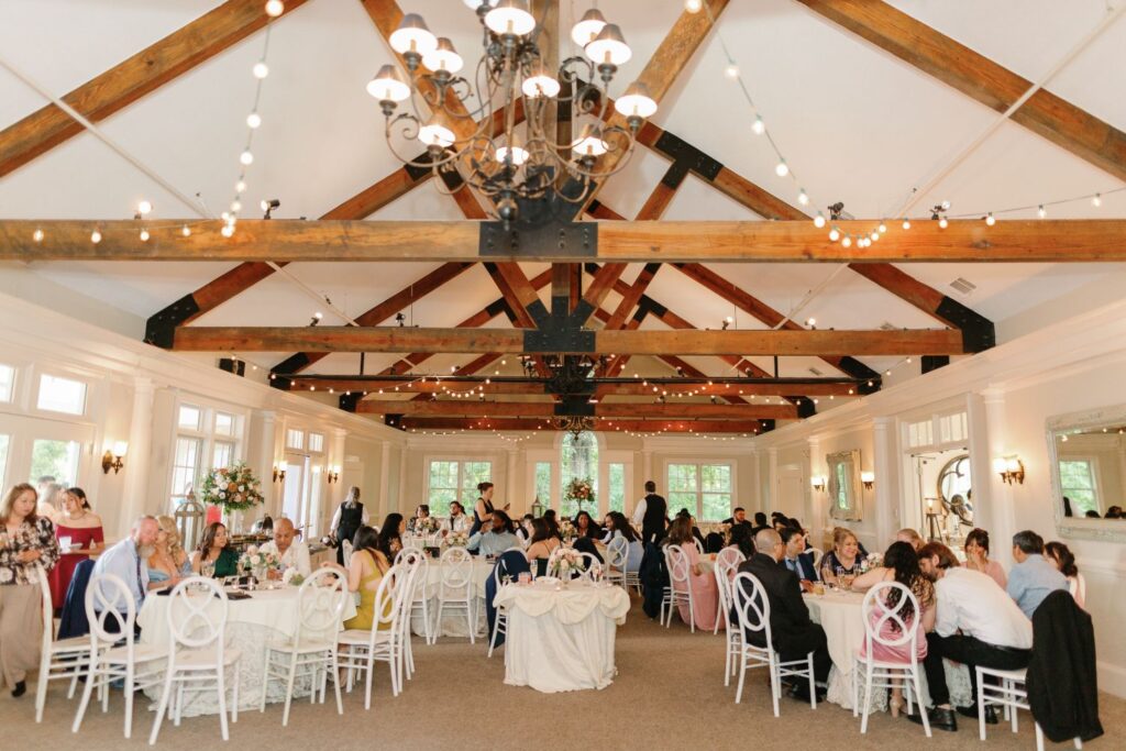 A large, elegant banquet hall with exposed wooden beams and string lights hosts a progressive cocktail reception Atlanta-style, as guests dine, converse, and mingle at round tables in the bright, airy space.