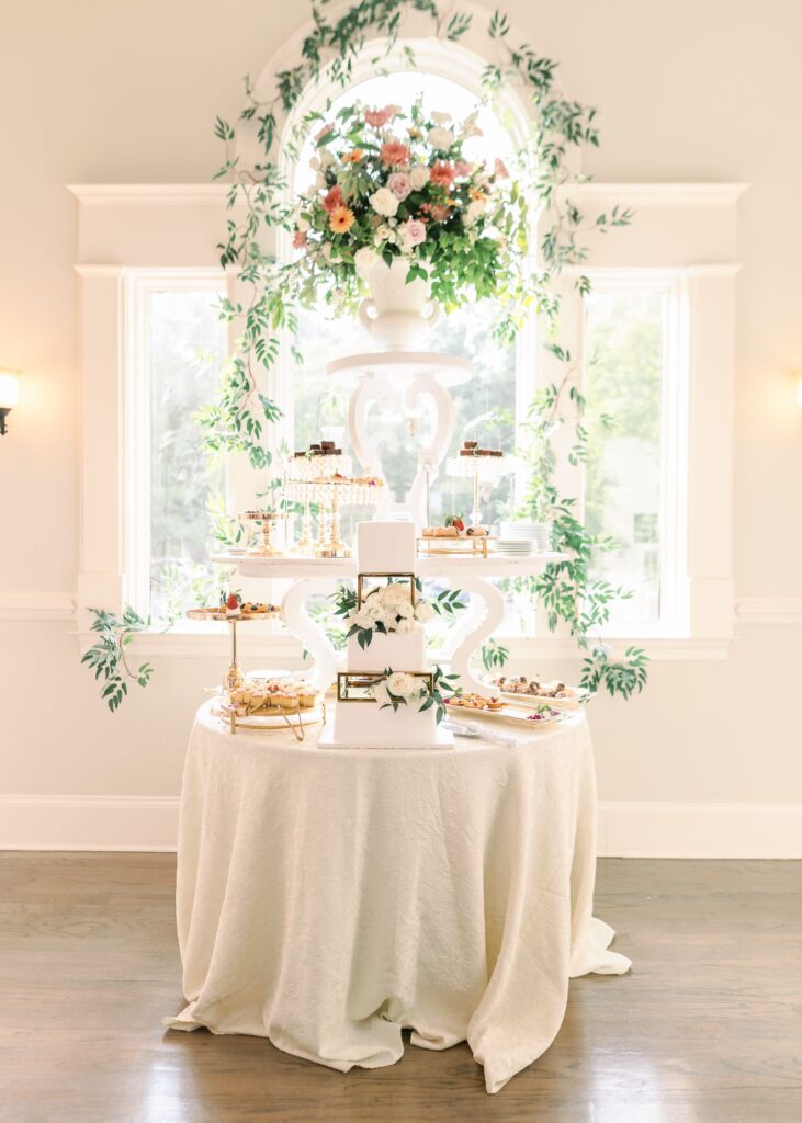 An elegant dessert table covered with a white tablecloth, featuring a floral cake, assorted pastries on gold stands, and floral arrangements—perfect for a progressive cocktail reception Atlanta style—in front of a bright window draped with greenery.