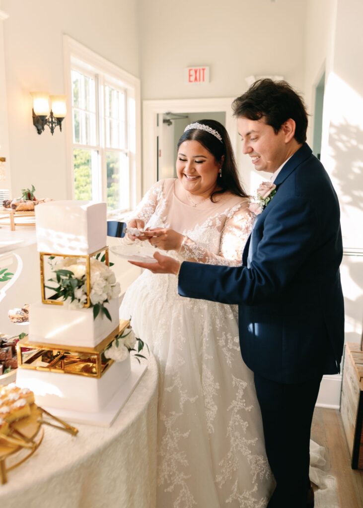 A bride in a lace gown and a groom in a navy suit smile as they cut a white tiered wedding cake with gold accents together during their progressive cocktail reception in sunlit Atlanta.