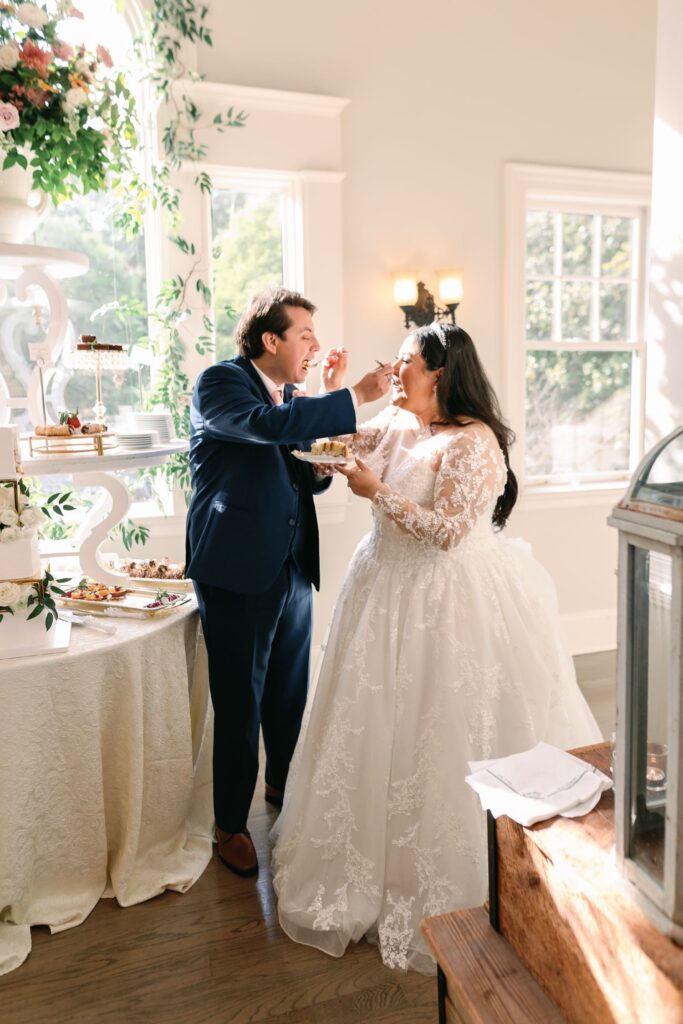 A bride and groom in wedding attire laugh and feed each other cake near a decorated dessert table during a progressive cocktail reception in a sunlit Atlanta room with large windows.