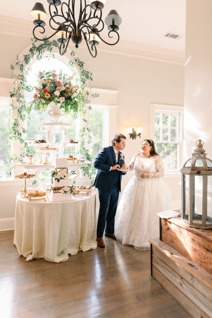 A bride and groom stand by a dessert table decorated with flowers in a sunlit room during their progressive cocktail reception in Atlanta. The groom holds a plate and chats with the bride, who wears a long-sleeved white wedding dress.