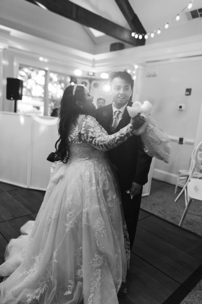 A bride in a lace wedding dress and a groom in a suit share a dance at their progressive cocktail reception in Atlanta, smiling and holding hands under string lights in the beautifully decorated indoor venue.