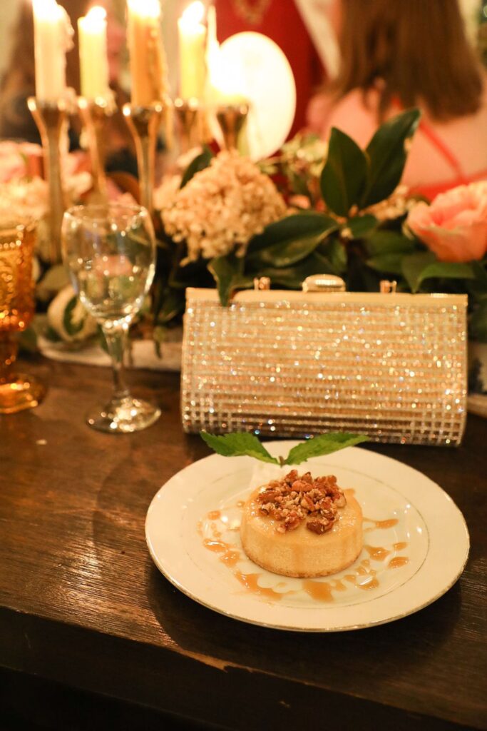 A dessert topped with nuts and caramel sauce sits on a plate at a Chastain Horse Park rehearsal dinner, in front of a sparkly clutch purse, with candles, flowers, a wine glass, and blurred people in the background on a wooden table.