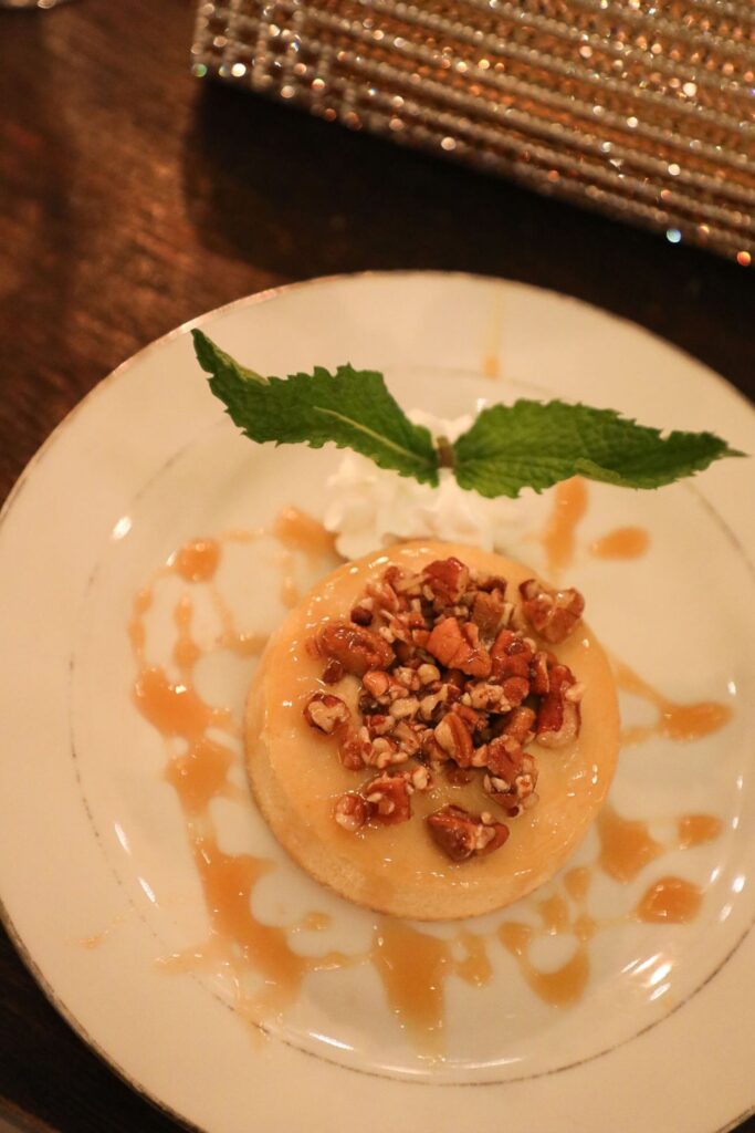 A round dessert topped with chopped nuts and caramel sauce sits on a white plate, garnished with whipped cream and two mint leaves—an elegant treat perfect for a Chastain Horse Park rehearsal dinner on a dark wooden table.