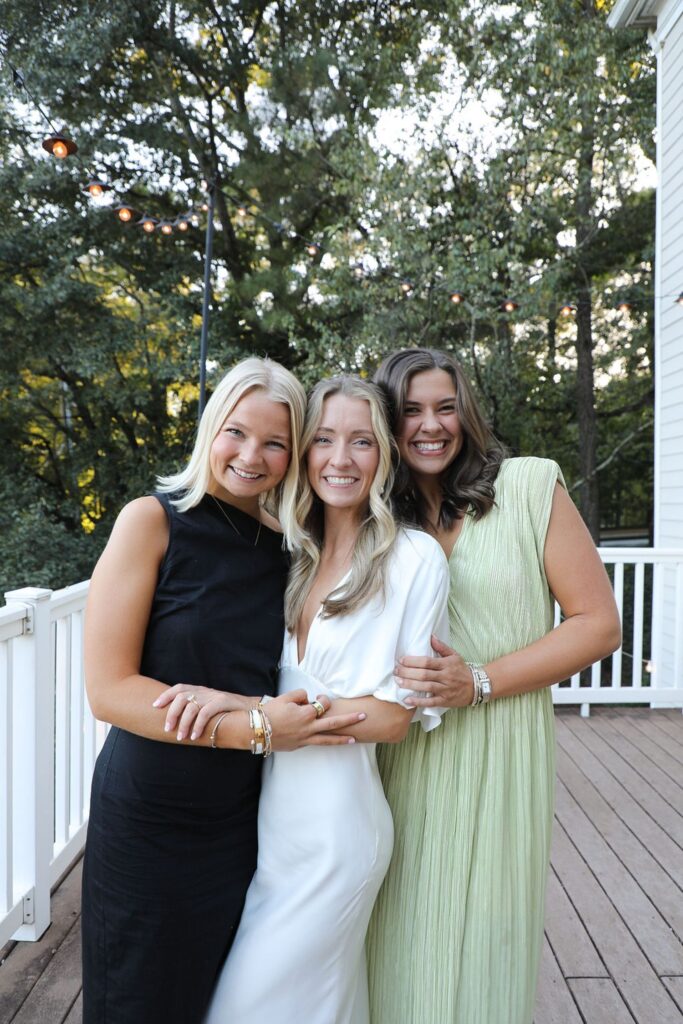 Three smiling women stand close together on a wooden deck at a Chastain Horse Park rehearsal dinner. They wear elegant dresses—black, white, and light green—surrounded by trees and glowing string lights in the background.