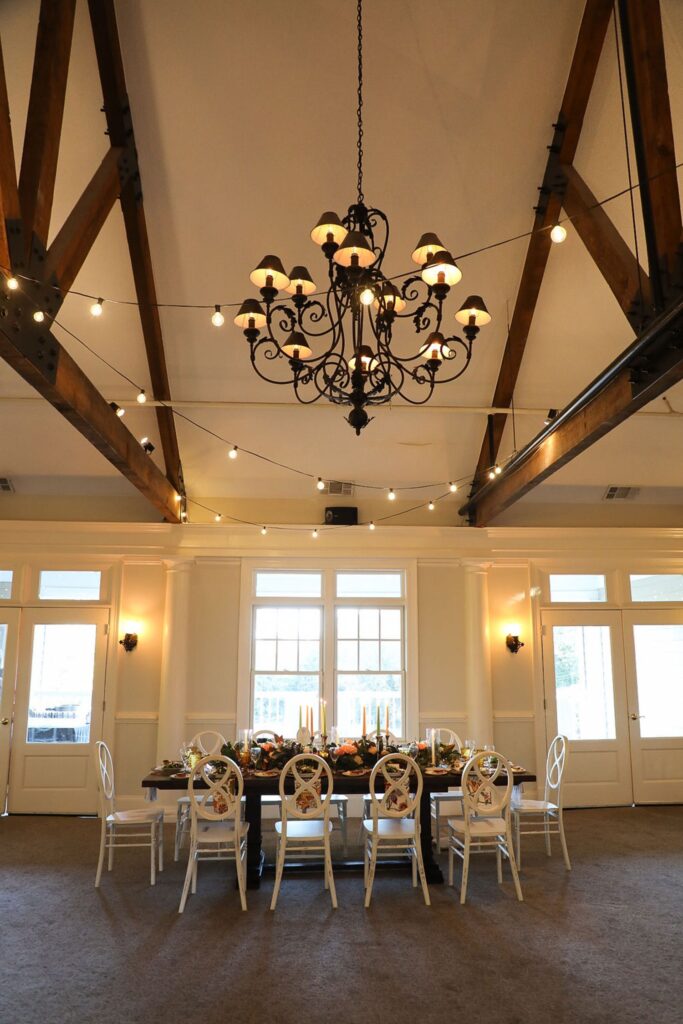 A large dining table set for a meal sits below a black chandelier with exposed wooden beams and string lights, creating an elegant setting reminiscent of a Chastain Horse Park rehearsal dinner in a bright room with white walls and tall windows.
