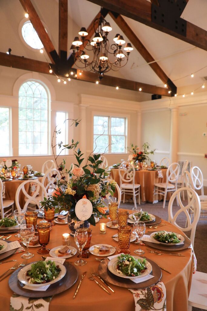 Elegant dining room set for a Chastain Horse Park rehearsal dinner with round tables, tan tablecloths, white chairs, floral centerpieces, salads on plates, amber glassware, and twinkling string lights under wooden beams.