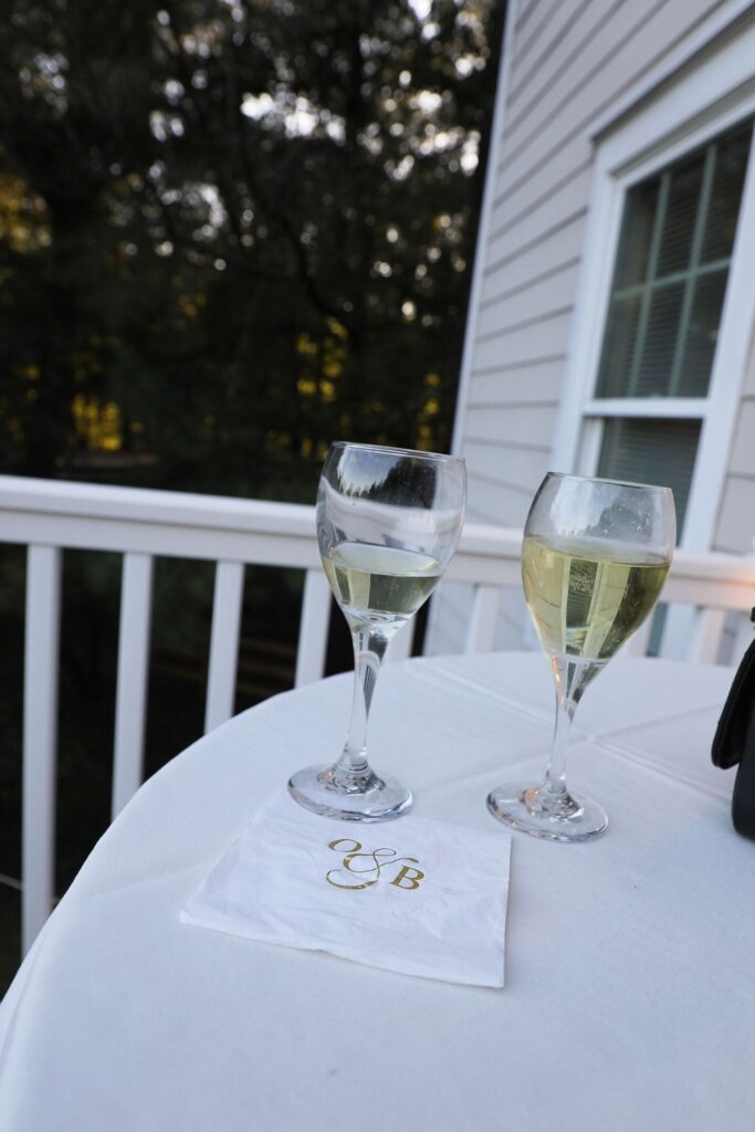 Two wine glasses with white wine sit on a round white table outside near a white railing at a Chastain Horse Park rehearsal dinner. A white napkin with gold initials G & B is placed on the table, with a building and siding in the background.
