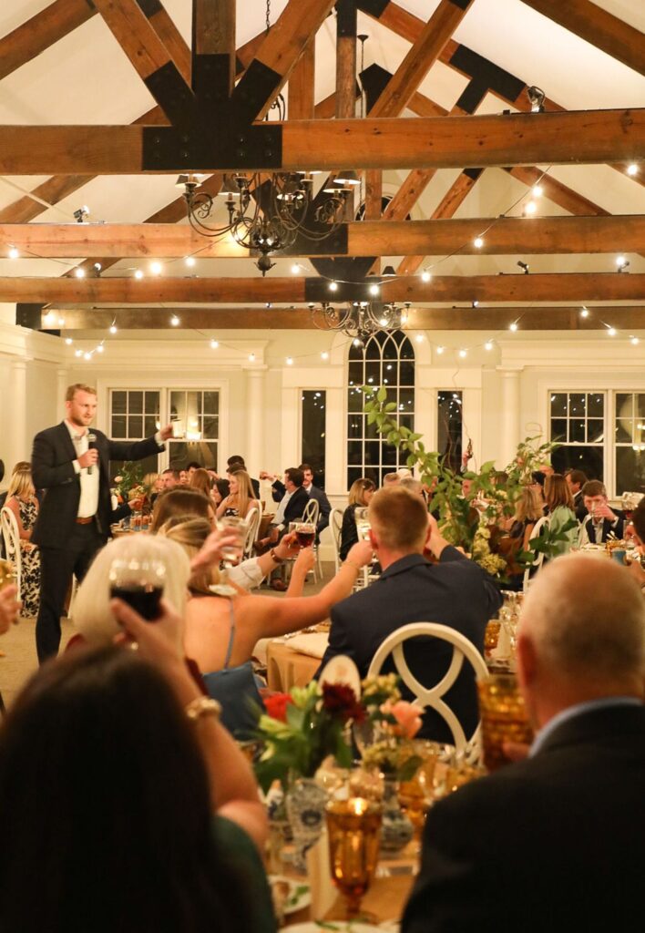 A man in a suit gives a toast with a microphone to a seated crowd at a Chastain Horse Park rehearsal dinner, with string lights, exposed wooden beams, and decorated tables creating an elegant indoor ambiance.