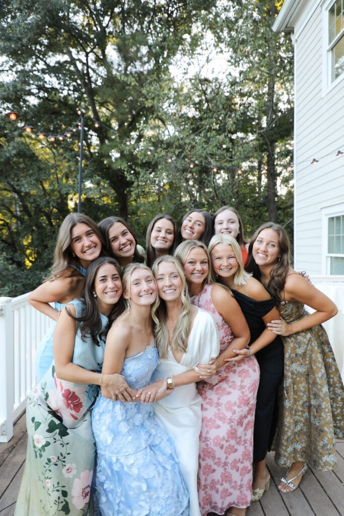 A group of twelve young women in colorful formal dresses smile and pose closely together on a wooden deck at a Chastain Horse Park rehearsal dinner, with trees and a white house in the background.
