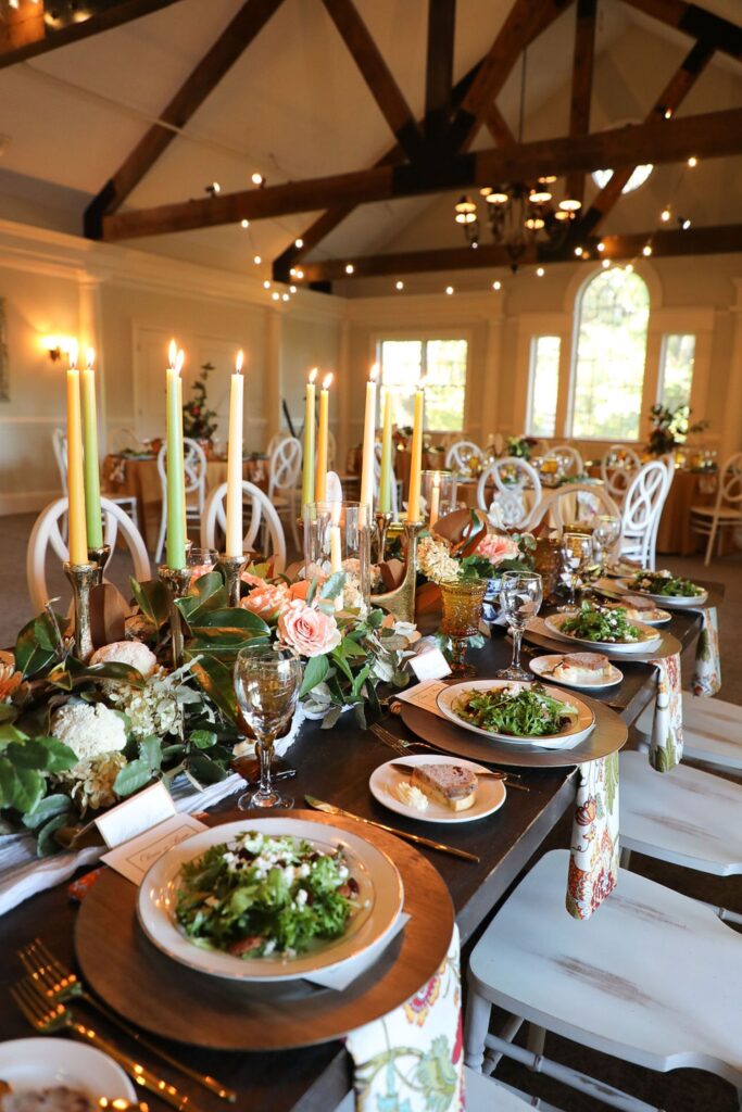 A long, elegant dining table set for a Chastain Horse Park rehearsal dinner features plates of salad, bread, wine glasses, gold candlesticks with tall yellow candles, floral centerpieces, and white chairs in a warmly lit, decorated room.