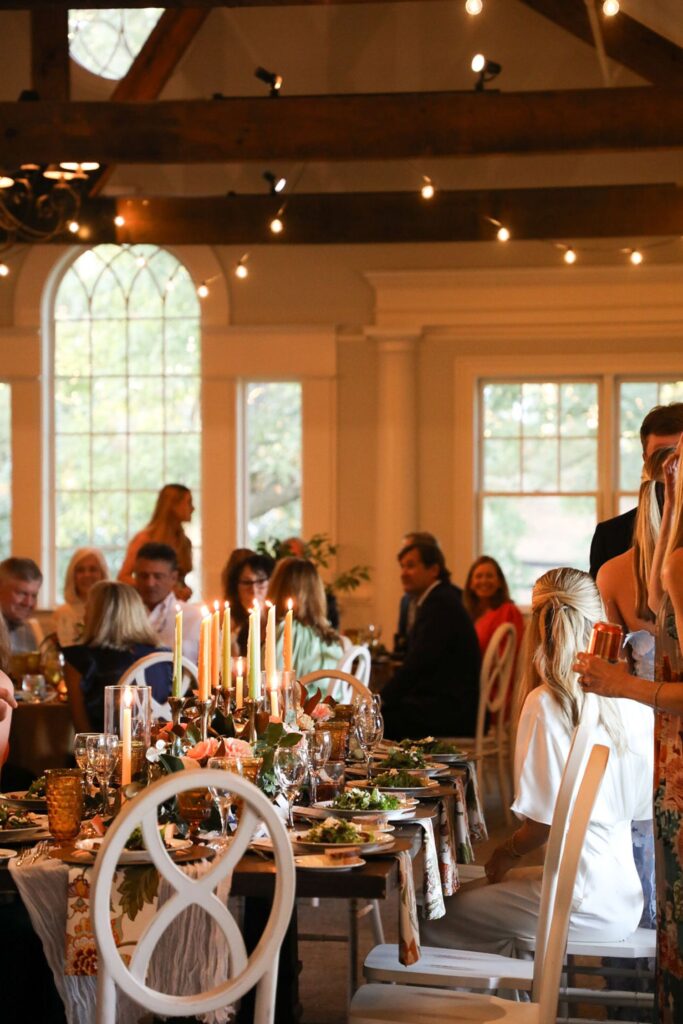 A group of people sit at decorated tables with candles, greenery, and food in a warmly lit room with large windows, wooden beams, and string lights, suggesting a festive Chastain Horse Park rehearsal dinner.