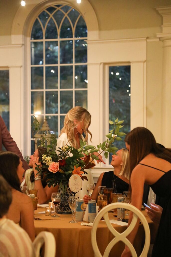 A group of women gather around a decorated table at a Chastain Horse Park rehearsal dinner, with floral centerpieces, drinks, and candles. One woman in white stands and chats with a seated woman in black. Large arched windows frame the background.