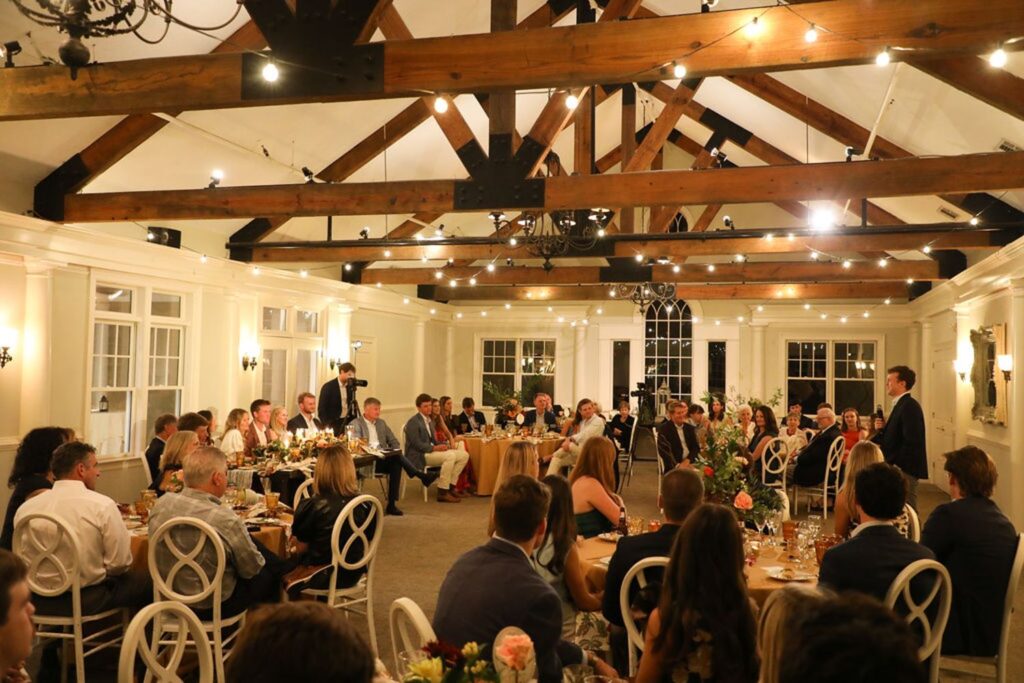 A large group of people seated at round tables in a decorated hall with exposed wooden beams at Chastain Horse Park, listening to a speaker; the atmosphere is formal and festive with string lights overhead, perfect for a rehearsal dinner.