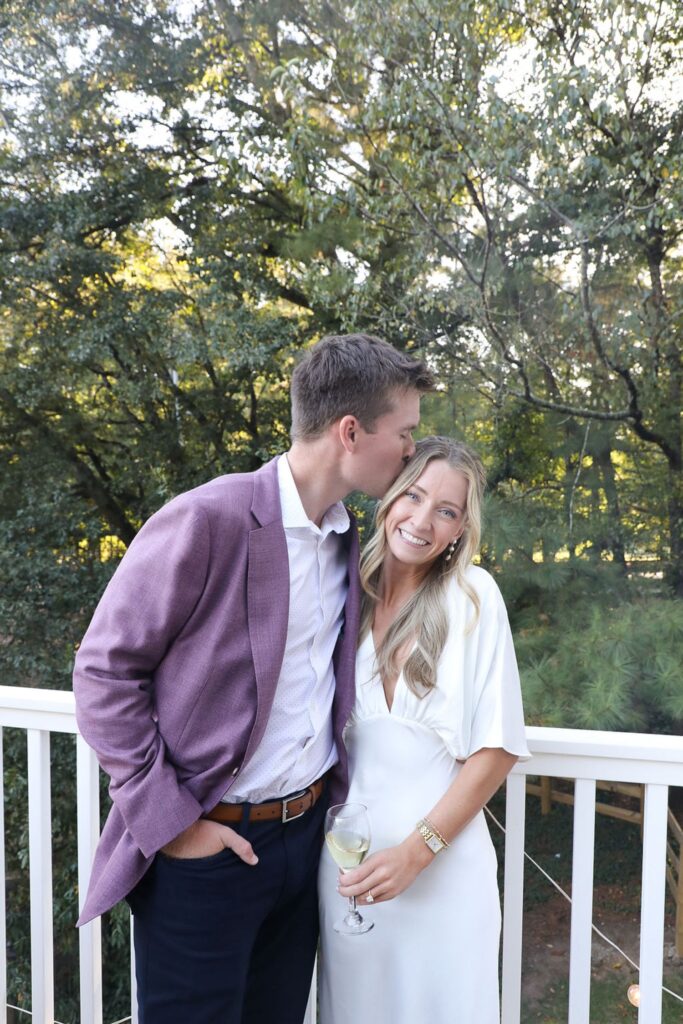 A man in a purple jacket kisses the forehead of a smiling woman in a white dress as they stand together on a balcony at their Chastain Horse Park rehearsal dinner, surrounded by green trees. She holds a glass of white wine.