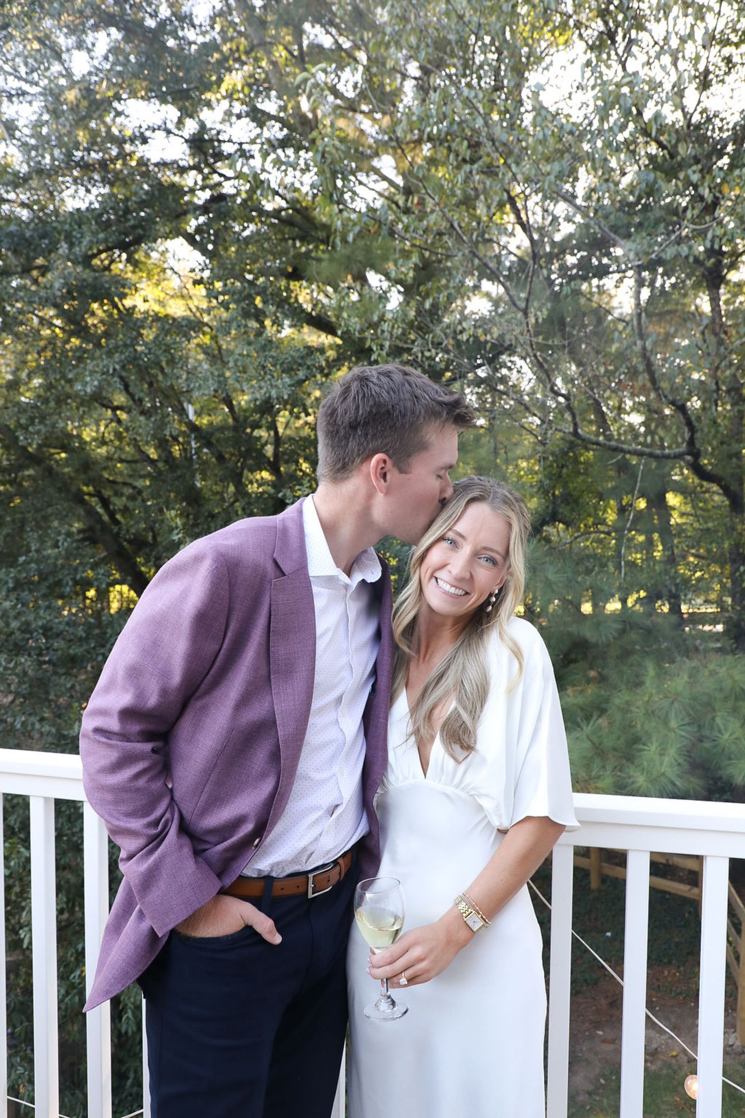 A man in a purple jacket kisses the forehead of a smiling woman in a white dress as they stand together on a balcony at their Chastain Horse Park rehearsal dinner, surrounded by green trees. She holds a glass of white wine.