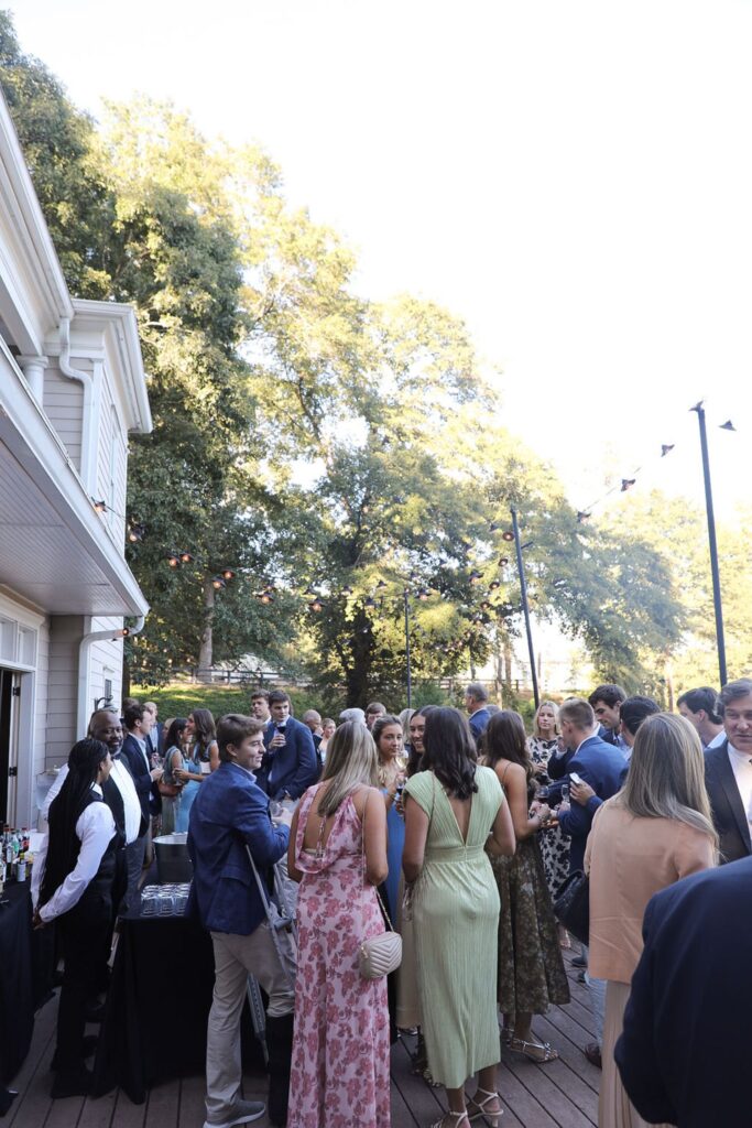 A group dressed in semi-formal attire mingle on a sunlit outdoor deck at Chastain Horse Park for a rehearsal dinner. With string lights overhead and trees nearby, guests gather by a white building as a bartender stands ready with drinks.