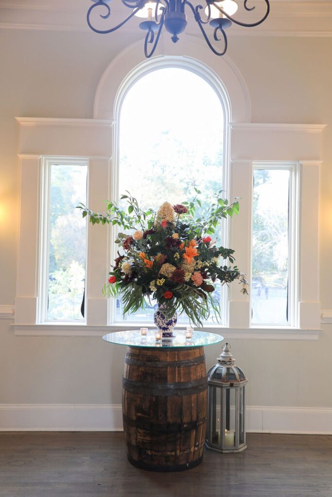 A floral arrangement in a blue and white vase sits on a glass-topped wooden barrel in front of tall windows, with a large lantern and small candles nearby, setting an elegant scene for a Chastain Horse Park rehearsal dinner.