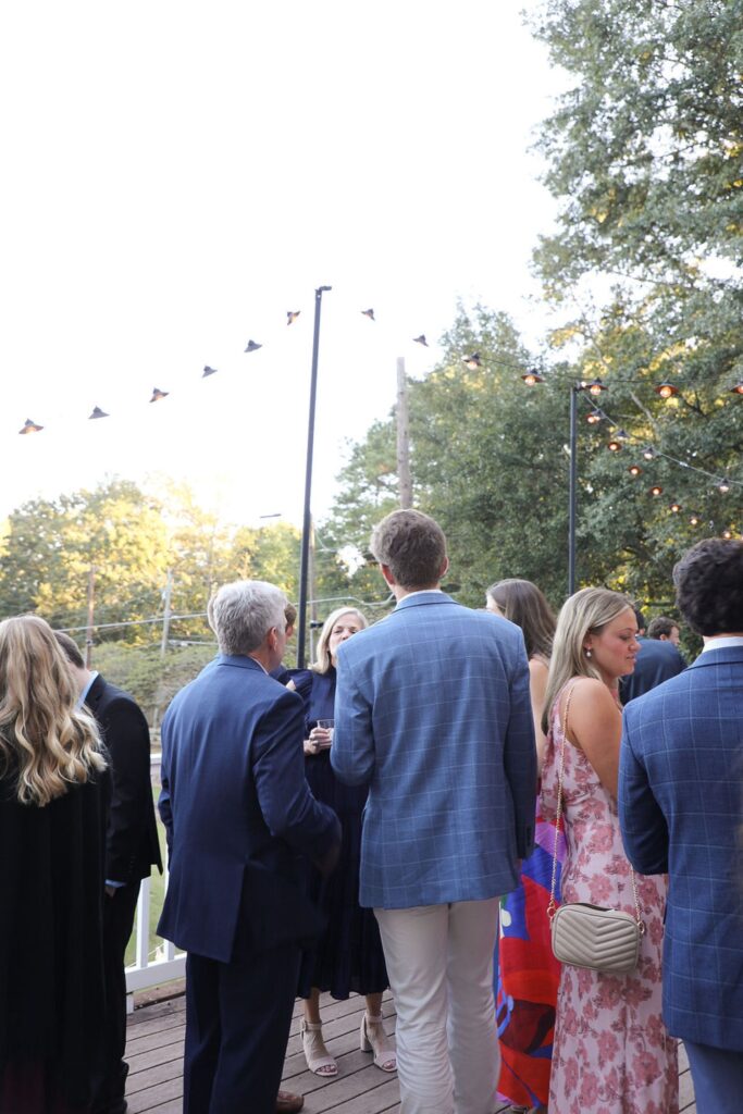 A group of people dressed in semi-formal attire stand and converse outdoors on a wooden deck at a Chastain Horse Park rehearsal dinner, surrounded by trees and string lights in the early evening.