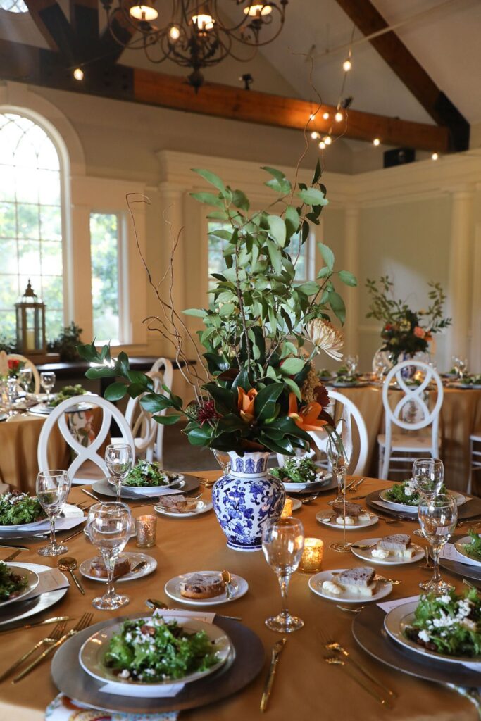 A formal dining table set for a meal, perfect for a Chastain Horse Park rehearsal dinner, with a large blue and white floral vase centerpiece, greenery, gold tablecloth, salad plates, wine glasses, and elegant place settings in a bright room.