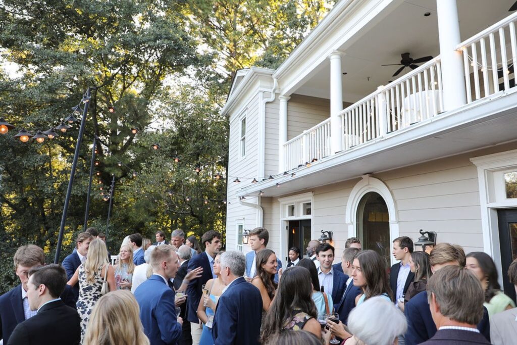 A group of well-dressed people mingle and converse outside a large, white house with a balcony at Chastain Horse Park, surrounded by trees and string lights on a sunny day, creating the perfect backdrop for a rehearsal dinner.