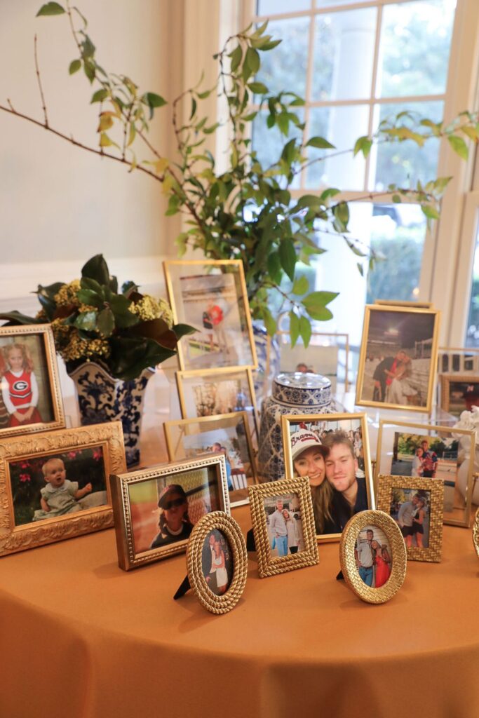 A display of framed family photos is arranged on a table with a gold tablecloth at the Chastain Horse Park rehearsal dinner. Greenery in vases and a large window in the background add natural light to the cozy scene.