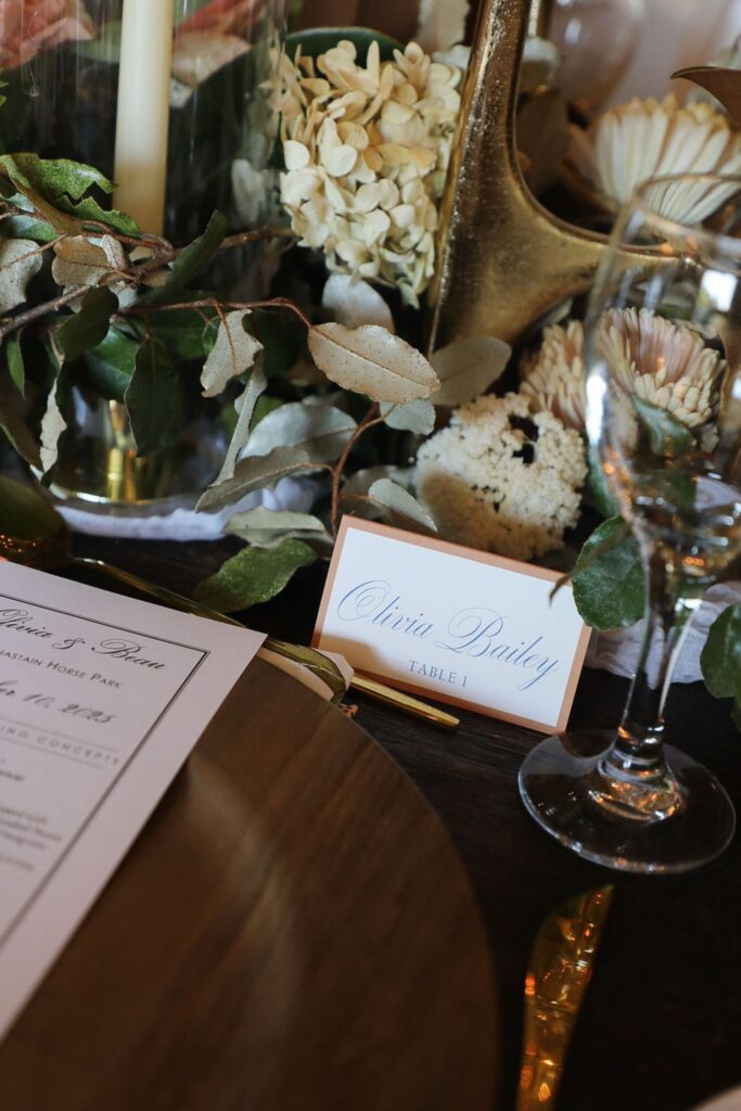 A close-up of an elegant table setting at a Chastain Horse Park rehearsal dinner features a place card reading Olivia Bailey, Table 1 amid greenery, flowers, a wine glass, and a menu on a wooden table.