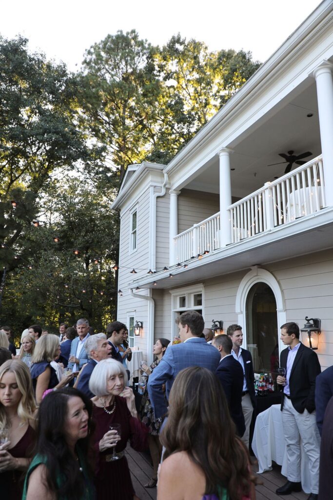 A crowd of people in semi-formal attire gather on a wooden deck outside a large, light-colored house with columns and a balcony, socializing during a daytime Chastain Horse Park rehearsal dinner event.