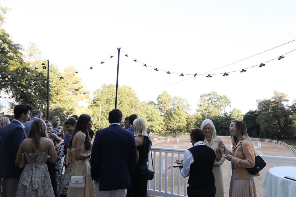 A group of people in semi-formal attire gather outdoors near a white fence at a Chastain Horse Park rehearsal dinner, socializing under string lights. Trees and an open field are visible in the background on a sunny day.