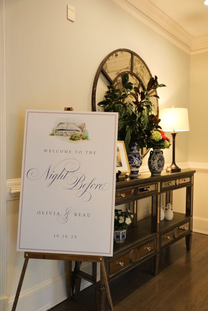 A decorative sign on an easel reads Welcome to The Night Before, Olivia & Beau, 10.10.25 in elegant script, setting the tone for a Chastain Horse Park rehearsal dinner in a warmly lit hallway with plants, a console table, and a mirror.
