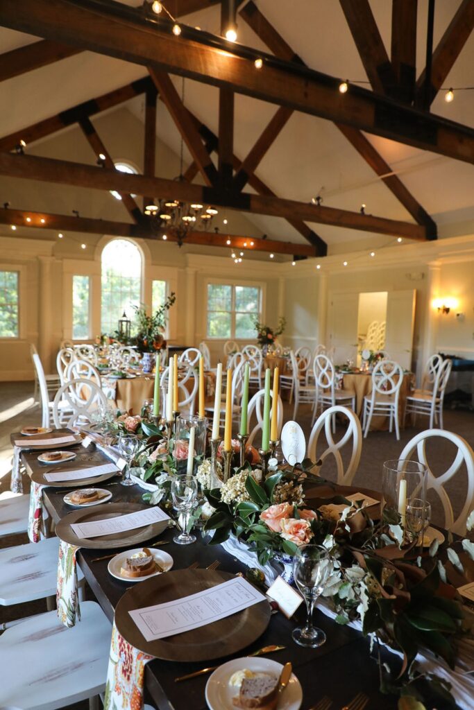 Elegant banquet hall set for a Chastain Horse Park rehearsal dinner with decorated tables, white chairs, greenery, candles, floral centerpieces, menus, and large windows letting natural light shine on exposed wooden beams above.