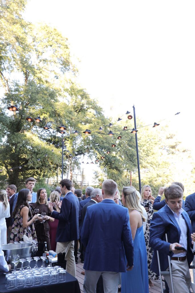 A group of well-dressed people gather outdoors under string lights for a Chastain Horse Park rehearsal dinner. Trees surround the area, and some guests hold drinks while conversing near a stylish bar setup with glassware in the foreground.