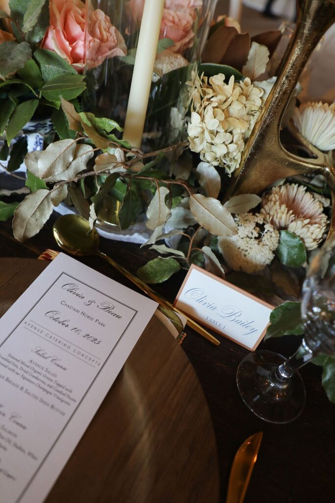 A close-up of an elegant table setting at a Chastain Horse Park rehearsal dinner, with a floral centerpiece, gold candle holder, menu, place card reading Olivia Bradley, wine glass, and gold cutlery on a wooden table.