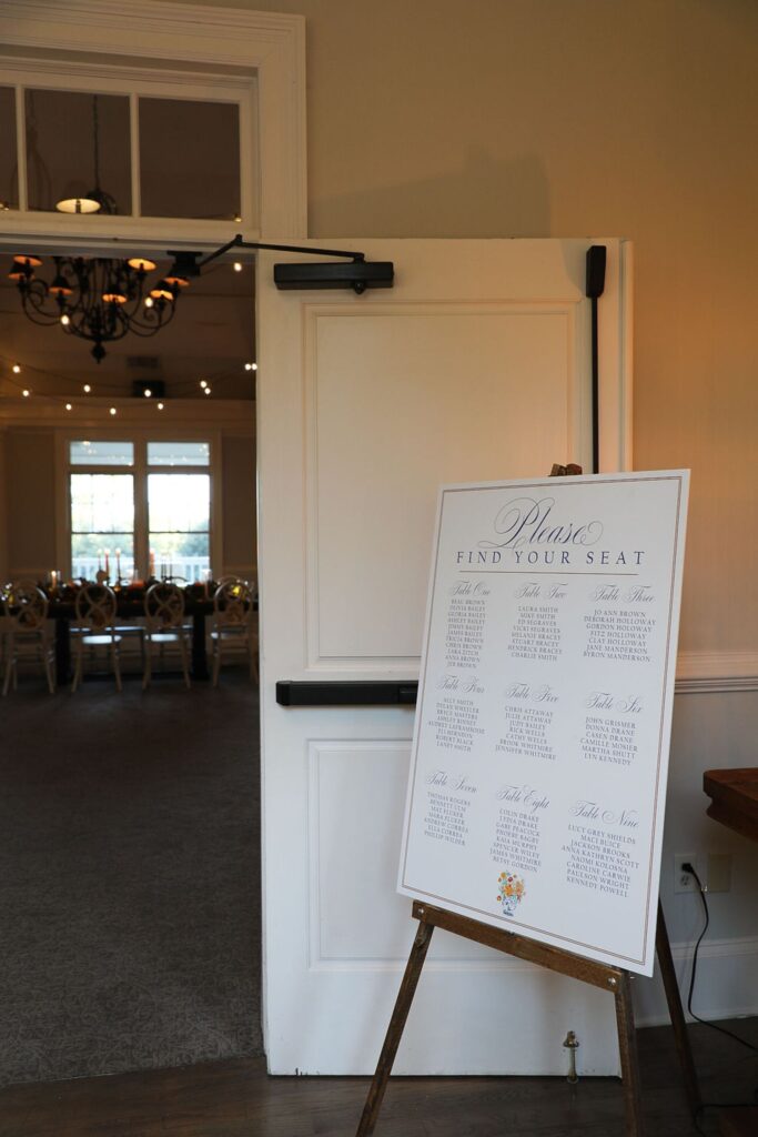 A white seating chart on an easel stands by the open door to a softly lit banquet hall at Chastain Horse Park, where round tables and chairs await guests for the rehearsal dinner under elegant chandeliers.