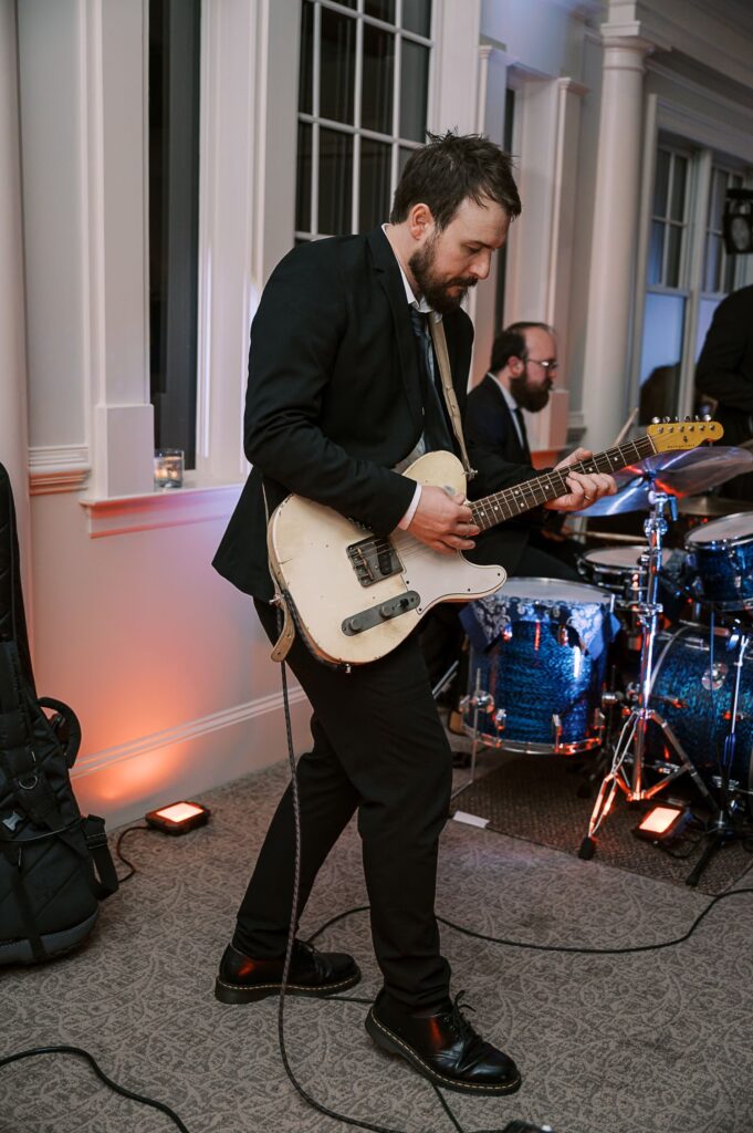 At a progressive buffet wedding in Atlanta, a man in a suit plays electric guitar next to a drum set in an indoor venue with large windows and beige carpet, while another musician on drums is visible in the background.