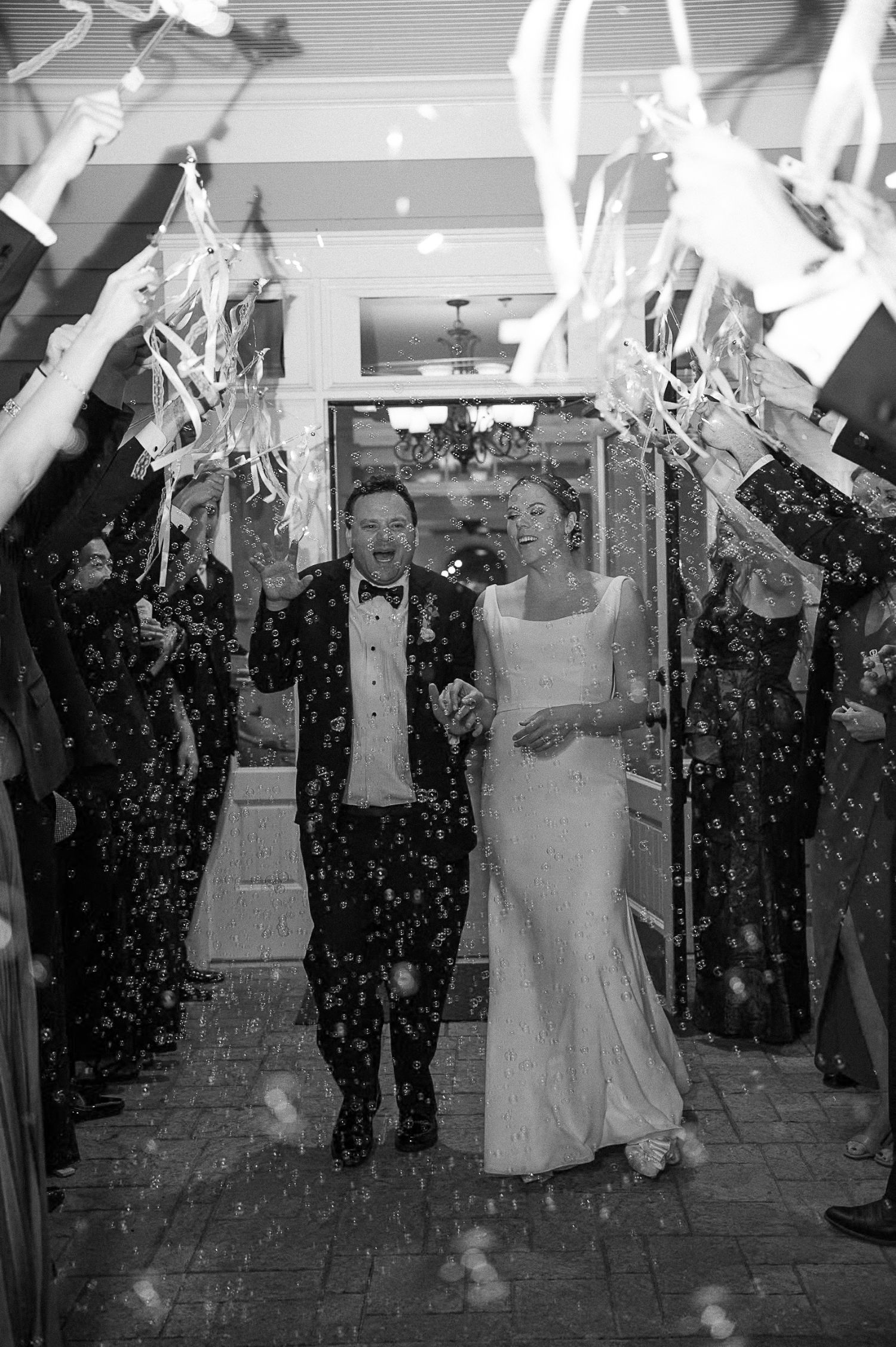 A bride and groom walk hand in hand through a corridor of guests waving ribbon wands and blowing bubbles at their progressive buffet wedding in Atlanta. Both are smiling and dressed formally; the scene is joyful and festive.