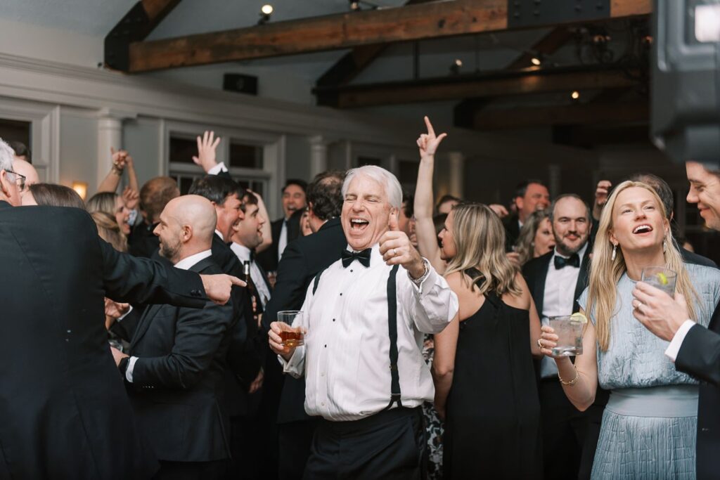 A joyful man in a tuxedo and suspenders dances and smiles at a crowded indoor progressive buffet wedding in Atlanta, holding a drink. Around him, people in formal attire are dancing, cheering, and enjoying the lively event.