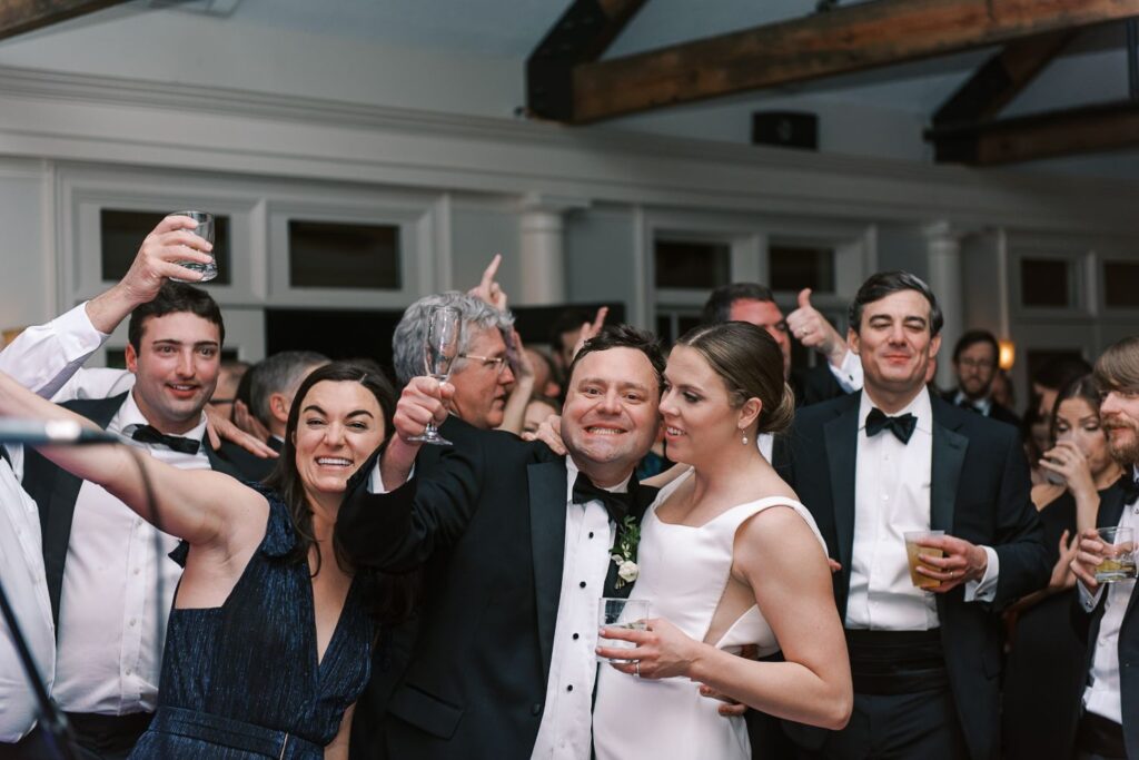 A group of well-dressed people at a progressive buffet wedding in Atlanta cheers and smiles for the camera. A couple in the center, the man in a tuxedo and the woman in a white dress, are surrounded by happy guests holding drinks.
