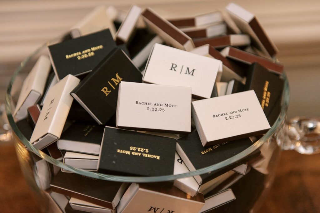 A glass bowl filled with personalized matchboxes in black, white, and brown, labeled “RIM” and “Rachel and Mote 2.22.25,” arranged as decorative wedding favors for a progressive buffet wedding in Atlanta.