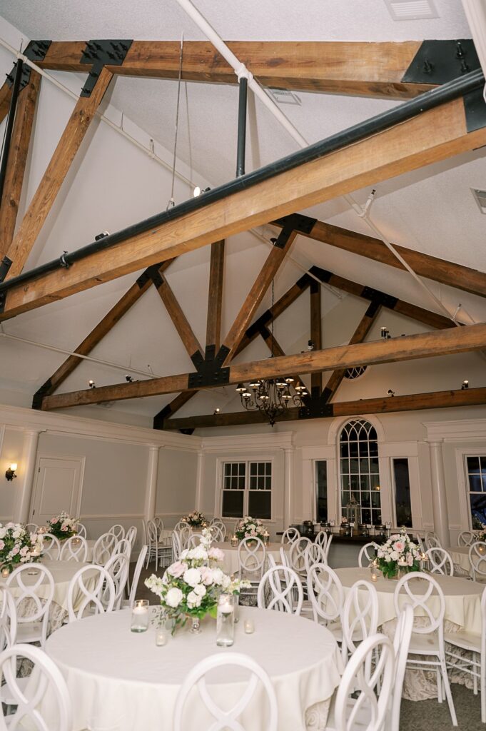 Elegant banquet hall with exposed wooden ceiling beams, round tables covered in white linens, and floral centerpieces—perfect for a progressive buffet wedding in Atlanta. Large windows and a chandelier enhance the sophisticated ambiance.