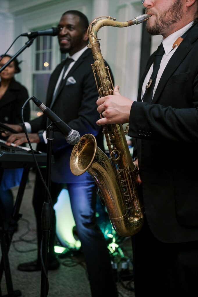 Two musicians perform indoors at a progressive buffet wedding in Atlanta; one plays saxophone while the other sings into a microphone and plays keyboard. Both are dressed in suits and ties.