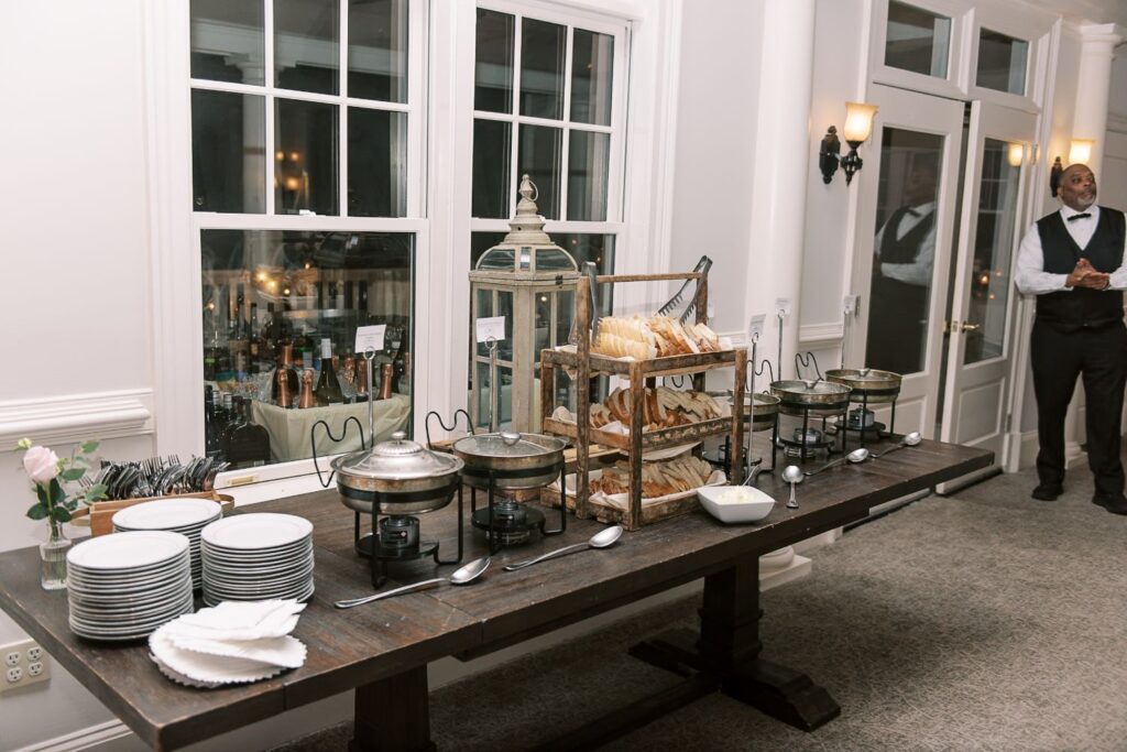 A buffet table set for a progressive buffet wedding in Atlanta features stacked plates, cutlery, chafing dishes, bread on tiered trays, and a lantern; a server in a vest and bow tie stands near the door in the well-lit room.