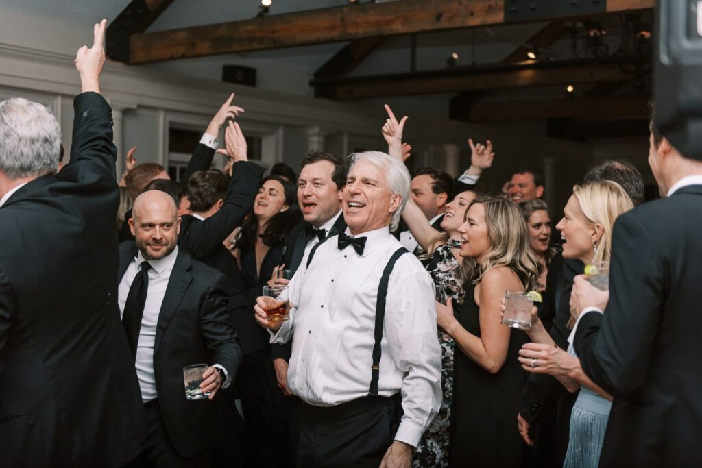 A group of well-dressed people, many in suits and dresses, celebrate and cheer together at a progressive buffet wedding in Atlanta. One older man in the front smiles widely, holding a drink, while others raise their hands and laugh.