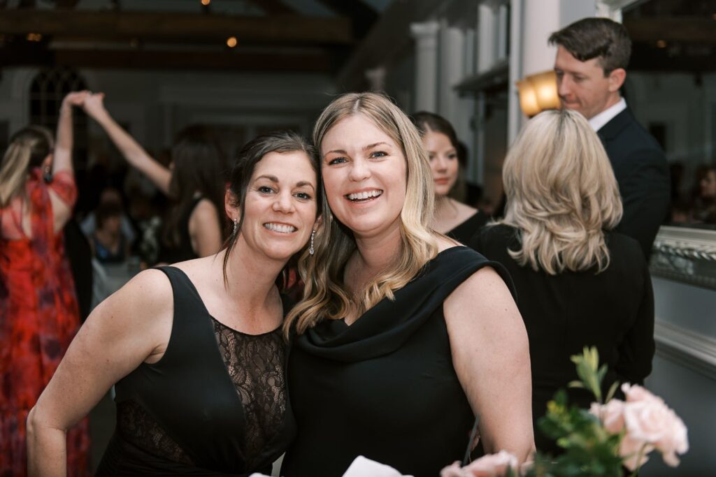 Two women smiling and posing together at a progressive buffet wedding in Atlanta. Both wear black dresses—one with lace detailing. In the background, guests are mingling and dancing, with a bouquet of pink flowers in the foreground.