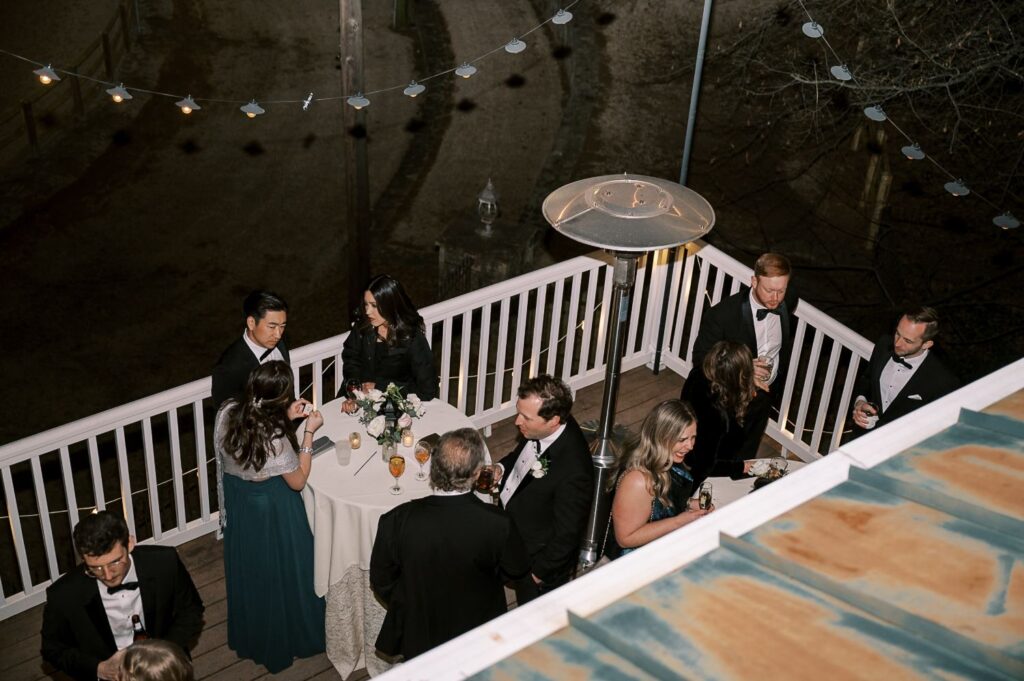A group of people in formal attire gather around a small round table on an outdoor deck at night during a progressive buffet wedding in Atlanta. String lights glow above, and a tall patio heater warms guests as they chat and enjoy drinks.